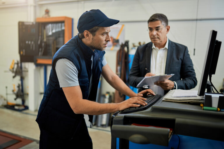 Mid adult inspector overseeing auto repairman who is working n a computer in a workshop. Focus is on mechanic.